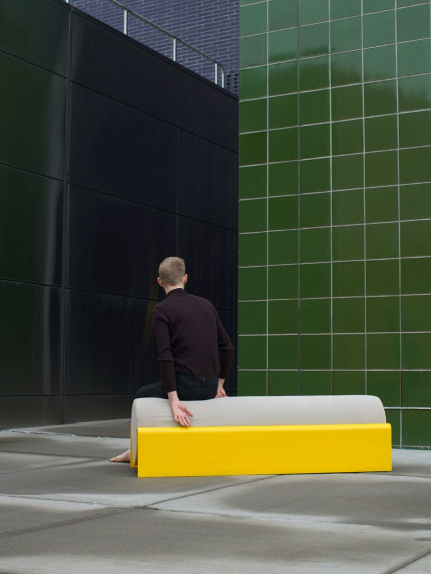 Man sitting on A modern minimal bench with a grey cushion and yellow fiberglass frame, outdoor furniture piece placed against a blue brick wall.