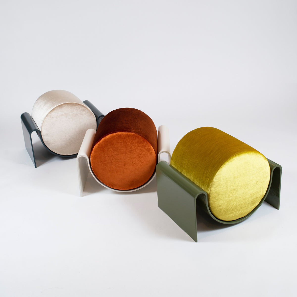 Three modern cylindrical side tables with sculptural U-shaped metal bases in black, white, and olive green, holding round upholstered tops in cream, rust orange, and mustard yellow, arranged on a light gray studio background.