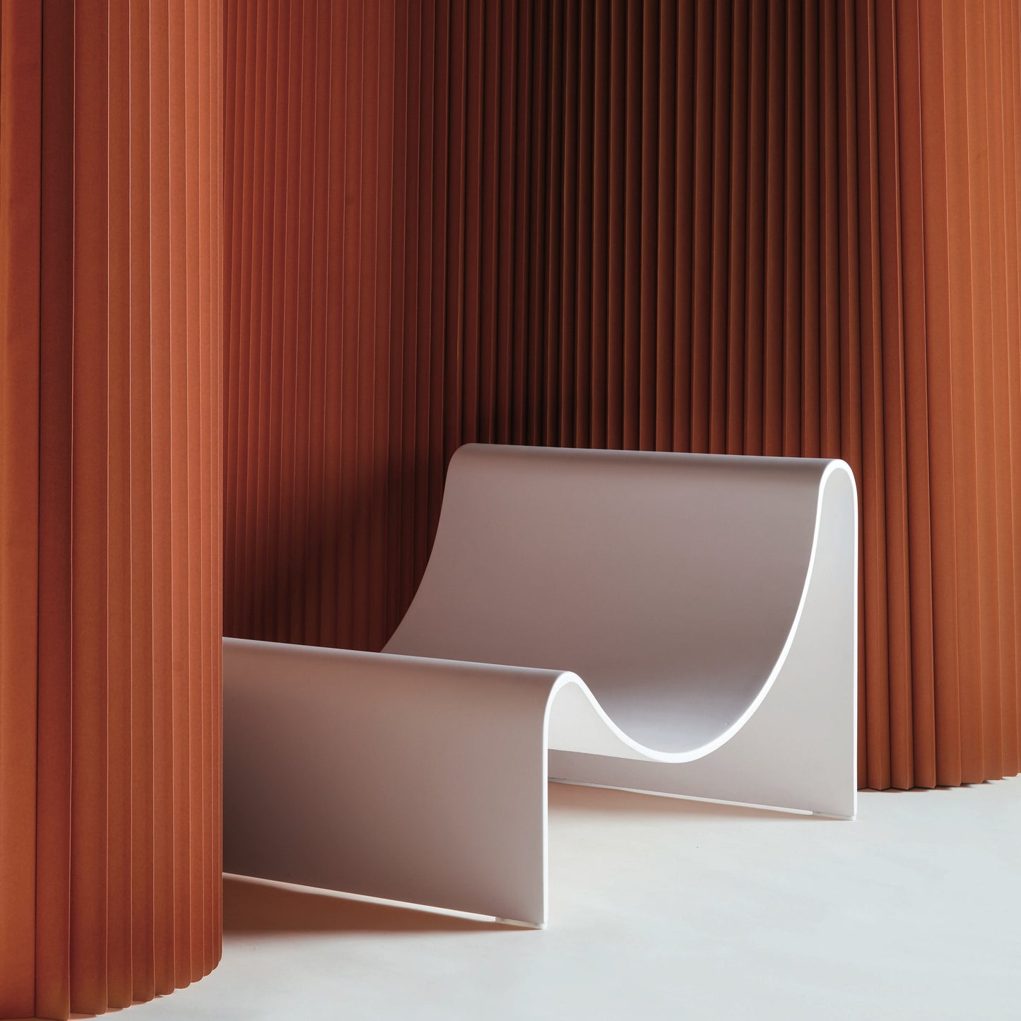 A modern, sleek lounge chair with a smooth, curved design, made of fiberglass, positioned indoors with a pleated orange curtain in the background.